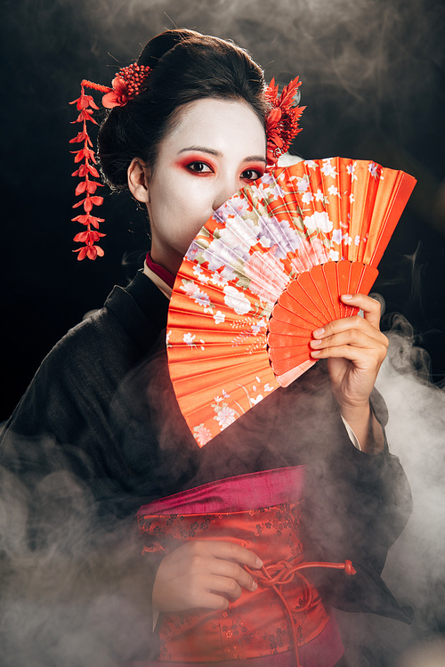 beautiful geisha in black kimono with flowers in hair holding hand fan in smoke