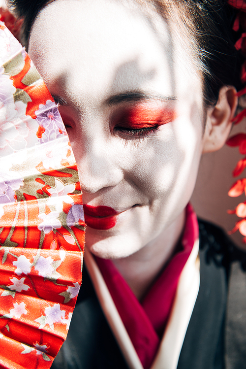 portrait of smiling beautiful geisha with red and white makeup holding hand fan in sunlight