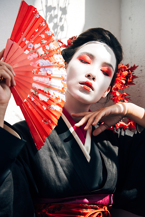 portrait of beautiful geisha with red and white makeup holding hand fan and touching face in sunlight