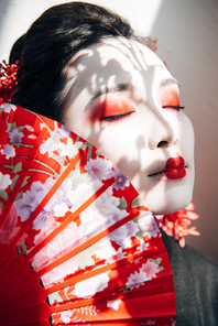 portrait of beautiful geisha with red and white makeup and closed eyes holding hand fan in sunlight