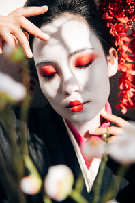 selective focus of tree branches and beautiful geisha with red and white makeup and closed eyes in sunlight