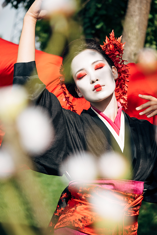 selective focus of tree branches and beautiful dancing geisha with red cloth on background in sunlight
