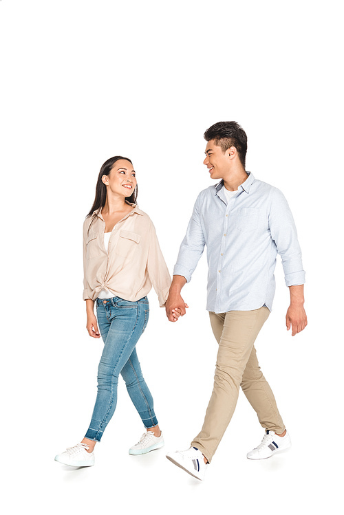 happy asian couple holding hands and looking at each other while walking on white background
