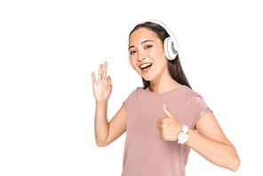 pretty asian woman listening music in headphones, showing thumb up and okay gesture while smiling at camera isolated on white