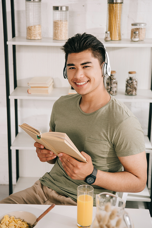 cheerful asian man in headphones smiling at camera while sitting at kitchen table and holding book