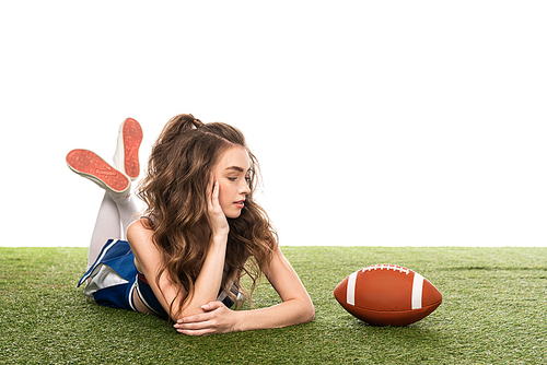 cheerleader girl in blue uniform lying near rugby ball on green field isolated on white