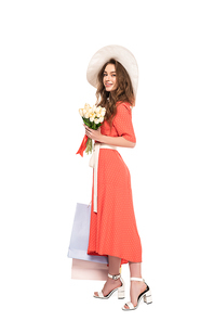 happy elegant woman in hat and dress holding white tulips and shopping bags isolated on white