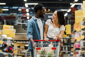 happy african american man looking at asian girl gesturing near shopping cart in supermarket