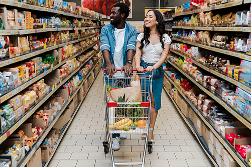 happy asian woman and african american man walking with shopping cart in supermarket