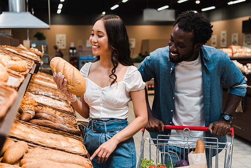 cheerful asian woman holding bread near african american man standing with shopping cart
