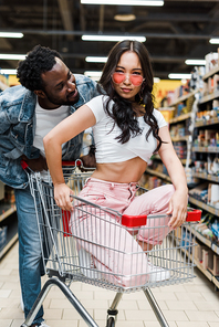 handsome african american man looking at stylish asian girl in sunglasses sitting in shopping cart in store