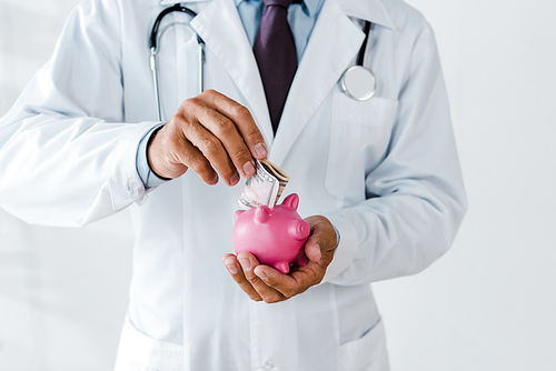 cropped view of man in white coat putting dollar banknote while holding piggy bank on white