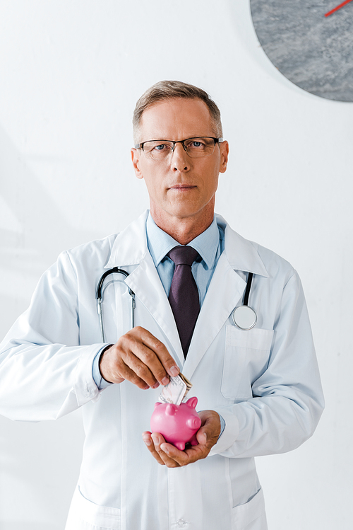 man in white coat putting dollar banknote while holding piggy bank