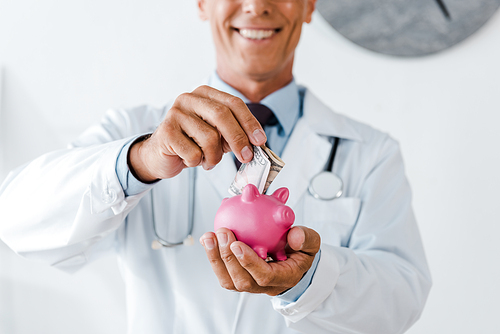 cropped view of cheerful man in white coat putting dollar banknote while holding piggy bank
