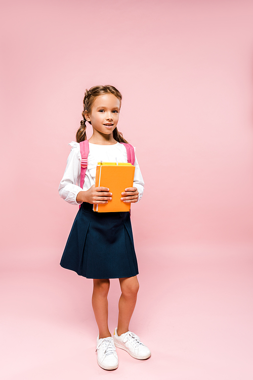 happy kid holding books while standing with backpack on pink