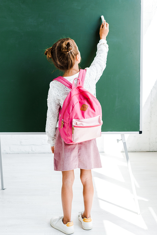 back view of schoolgirl standing with backpack and holding chalk near green chalkboard