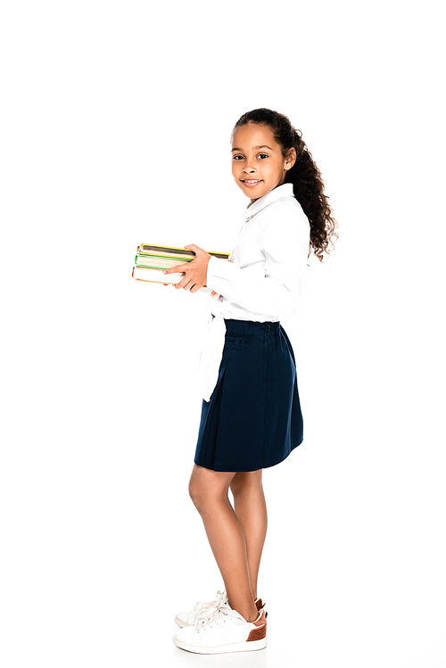 full length view of adorable african american schoolgirl holding books and smiling at camera on white background