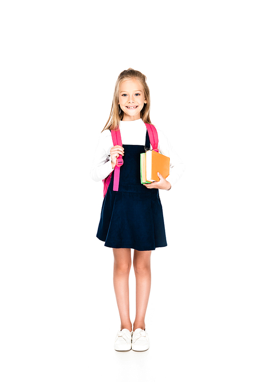 full length view of cute schoolgirl smiling at camera while holding books on white background