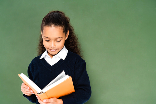 attentive african african american schoolgirl reading book while standing near chalkboard