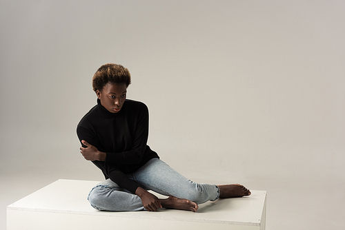 african american girl in black turtleneck sitting on white cube on grey