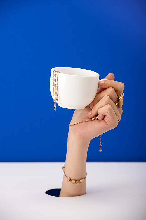 cropped view of woman with bracelet on hand holding cup with golden chain isolated on blue