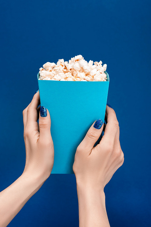 cropped view of woman holding box with popcorn isolated on blue