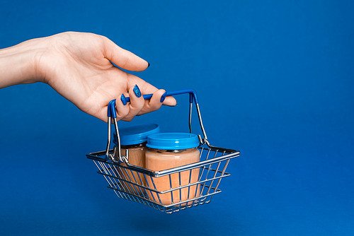 cropped view of woman holding shopping basket with baby food in jars on blue background
