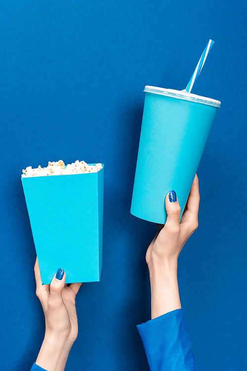 cropped view of woman holding popcorn and paper cup on blue background
