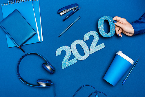 cropped view of woman holding number near notebooks, headphones, pens, stapler on blue background