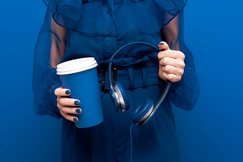 cropped view of woman holding paper cup and headphones on blue background