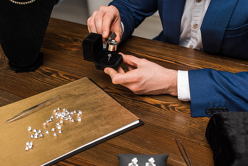 Cropped view of jewelry appraiser examining jewelry ring with magnifying glass near gemstones on wooden table