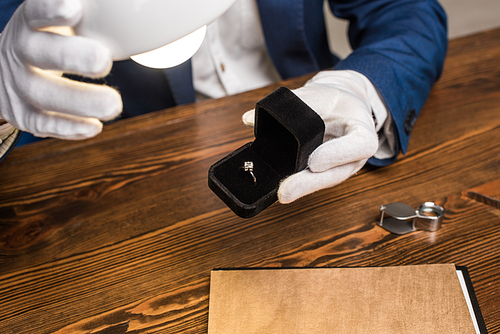 Cropped view of jewelry appraiser holding ring with gemstone in box near lamp at table isolated on grey