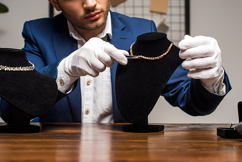 Cropped view of jewelry appraiser in gloves holding tweezers near necklace on necklace stand on table