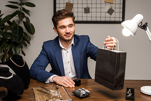 Handsome jewelry appraiser holding paper bag near earrings and necklaces on table