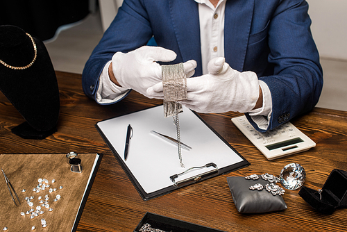 Cropped view of jewelry appraiser holding necklace near calculator, clipboard and jewelry on table