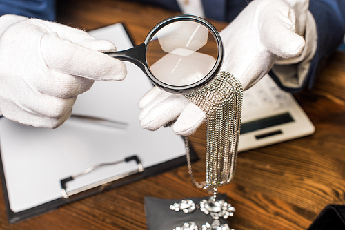 Cropped view of jewelry appraiser holding necklace and magnifying glass near calculator, clipboard and earrings on table