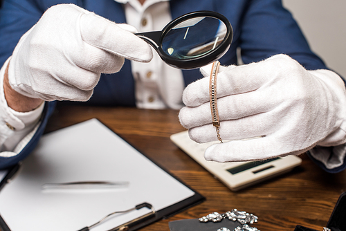 Cropped view of jewelry appraiser examining necklace with magnifying glass near calculator, clipboard and earnings on table isolated on grey