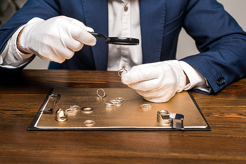 Cropped view of jewelry appraiser with magnifying glass examining jewelry ring near board on table on grey background