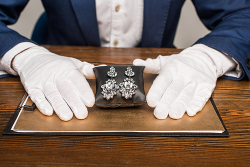 Cropped view of jewelry appraiser holding jewelry earnings on jewelry pillow near board on table isolated on grey
