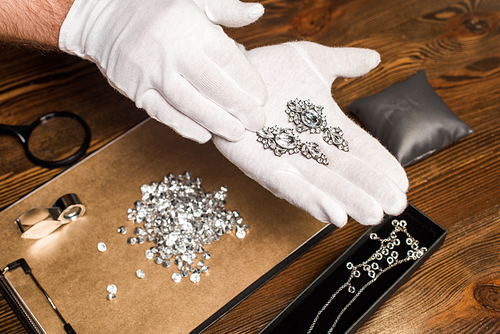 Cropped view of jewelry appraiser holding earrings near gemstones and magnifying glasses on board on table