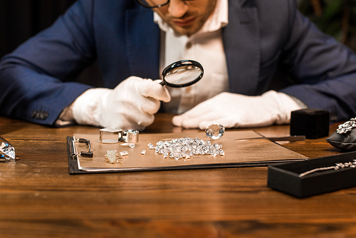 Cropped view of jewelry appraiser examining gemstones on board near jewelry on table isolated on black