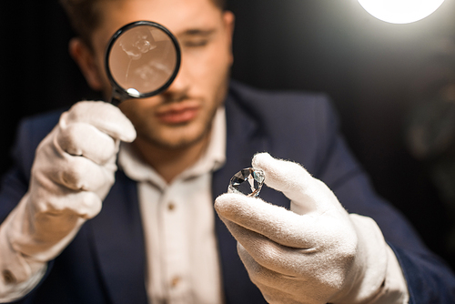 Selective focus of jewelry appraiser examining gemstone with magnifying glass on black background