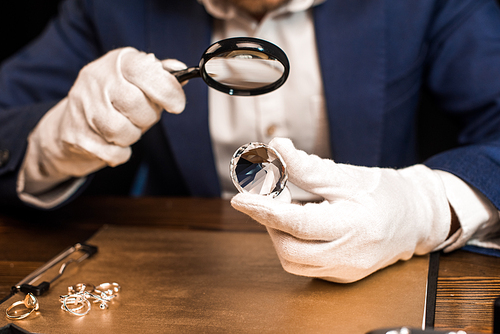 Cropped view of jewelry appraiser holding gemstone and magnifying glass near jewelry rings on board on table isolated on black