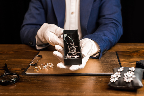 Cropped view of jewelry appraiser holding box with necklace near jewelry on board on table isolated on black