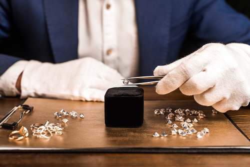 Cropped view of jewelry appraiser holding gemstone in tweezers near jewelry on board on table isolated on black