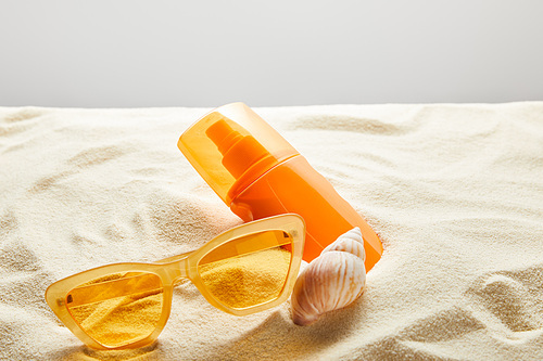 yellow stylish sunglasses and sunscreen in orange bottle on sand with seashell on grey background
