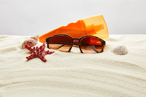 brown stylish sunglasses on sand with red starfish, seashells and sunscreen in orange bottle on grey background