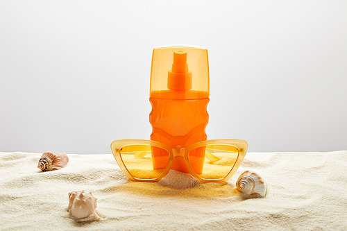 yellow stylish sunglasses and sunscreen in orange bottle on sand with seashells on grey background