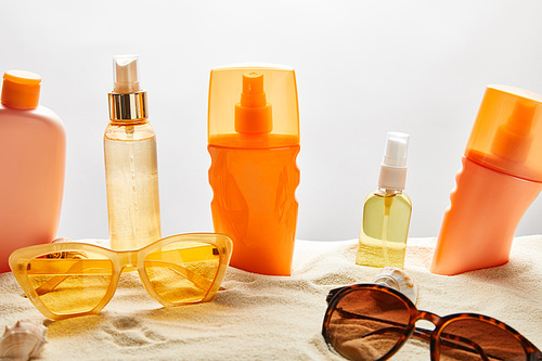 various sunscreen products in bottles on sand near fashionable sunglasses and seashells on grey background