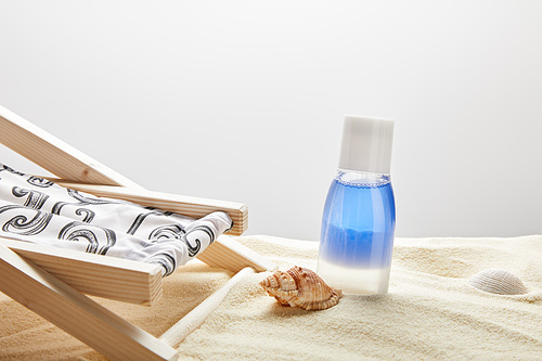 blue lotion in bottle in sand near seashells and deck chair on grey background with copy space